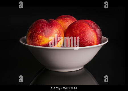 Group of three whole fresh red nectarine in a white ceramic bowl isolated on black glass Stock Photo