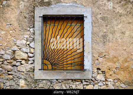 Old house window with metal bars in shape of morning sun closed shutter in textured rocks wall Stock Photo