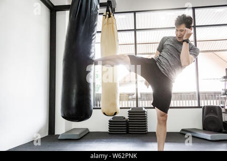 Young healthy fit Asian man practicing his kick with the punch sand bag at a local gym, modern lifestyle Stock Photo
