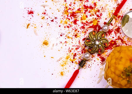 Indian Festival – Close view of elegant Rakhi, sweets, Cardamom and Indian currency with spread rice grains, kumkum, and turmeric on white background Stock Photo