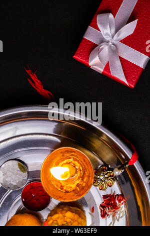 Indian Festival – Elegant Rakhi, rice grains, kumkum, sweets, diya in plate with red gift box on black surface. Festival of brother and sister Stock Photo