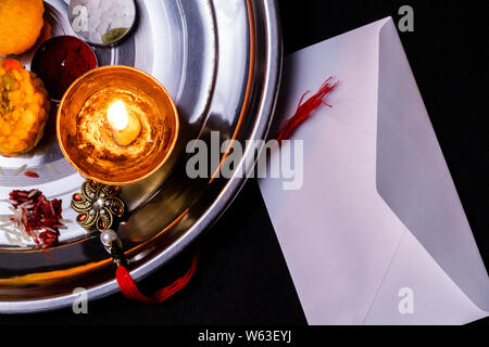 Raksha Bandhan - Elegant Rakhi, rice grains, kumkum, sweets, diya on plate with closed white envelope on dark background Stock Photo