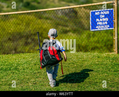 Young boy walking toward the baseball field carrying his bat and gear with fence in background. Stock Photo