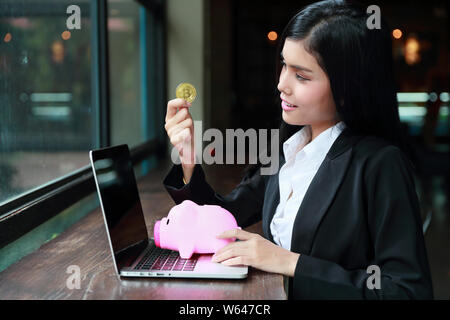 portrait of businesswoman showing bitcoin or coin on pink piggy with computer meaning of profit investment or money saving Stock Photo
