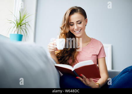 Joyful young woman smiling while reading book and holding coffee at home Stock Photo