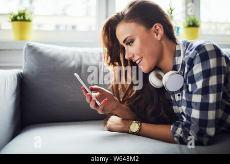 Side view of a young attractive woman smiling and looking at phone while lying on sofa Stock Photo