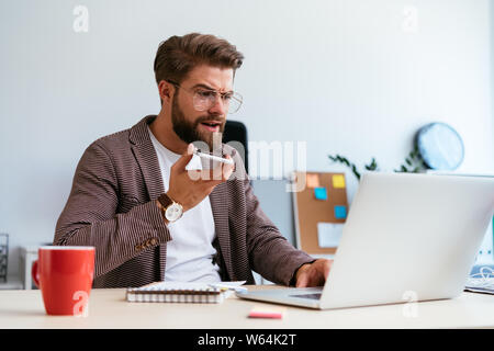 Concentrated startup businessman talking on phone with speaker mode while looking at laptop in office Stock Photo