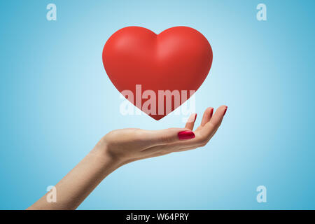 Side closeup of woman's hand facing up and levitating beautiful red heart on light-blue gradient background. Stock Photo