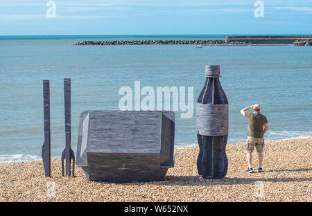 Lyme Regis, Dorset, UK. 31st July 2019. UK Weather: A man admires the view from a giant litter sculpture of takeaway packaging that has been erected on the picturesque beach at the seaside resort of Lyme Regis. Consisting of an 8 foot high plastic drinks bottle, cutlery measuring 6 foot tall and a chip/burger box measuring 4x4 foot the sculptures promote the efforts of Litter Free Coast and Sea and partners in promoting environmentally responsible choices. It is hoped that the temporary installation by architectural designer Jack Raisey will encourage people to take their litter home. Credit:  Stock Photo