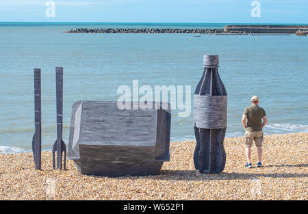 Lyme Regis, Dorset, UK. 31st July 2019. UK Weather: A man admires the view from a giant litter sculpture of takeaway packaging that has been erected on the picturesque beach at the seaside resort of Lyme Regis. Consisting of an 8 foot high plastic drinks bottle, cutlery measuring 6 foot tall and a chip/burger box measuring 4x4 foot the sculptures promote the efforts of Litter Free Coast and Sea and partners in promoting environmentally responsible choices. It is hoped that the temporary installation by architectural designer Jack Raisey will encourage people to take their litter home. Credit:  Stock Photo
