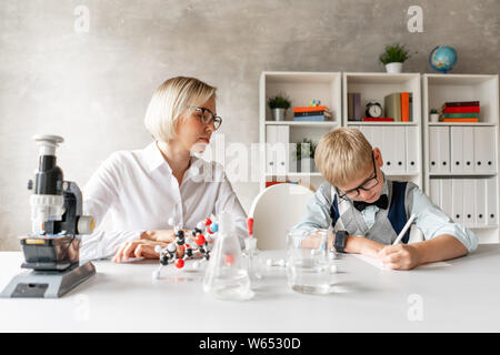 Blond schoolboy write properties of chemical element into his notebook under guidance of his charming teacher Stock Photo