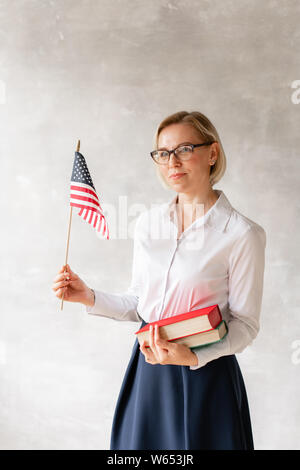 Charming teacher wearing blouse and skirt holds two books and flag in her hands Stock Photo