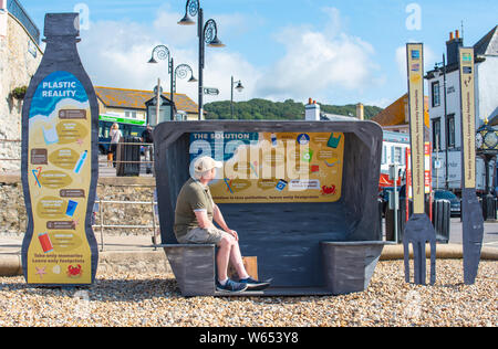 Lyme Regis, Dorset, UK. 31st July 2019. UK Weather: A man admires the view from a giant litter sculpture of takeaway packaging hat has been erected on the picturesque beach at the seaside resort of Lyme Regis. Consisting of an 8 foot high plastic drinks bottle, cutlery measuring 6 foot tall and a chip/burger box measuring 4x4 foot the sculptures promote the efforts of Litter Free Coast and Sea and partners in promoting environmentally responsible choices. It is hoped that the temporary installation by architectural designer Jack Raisey will encourage visitors to take their litter home. Credit: Stock Photo