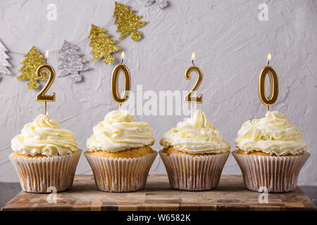 Golden candles in the form of figures 2020 on the cupcakes with white cream among the Christmas lights. New Year 2020 concept Stock Photo