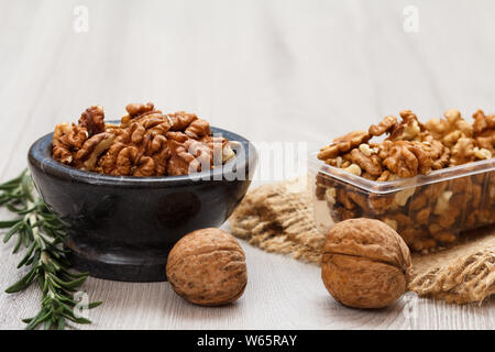 Peeled and unpeeled walnuts in stone bowl and plastic container with sackcloth and rosemary on a gray wooden background. Useful nutritious protein pro Stock Photo