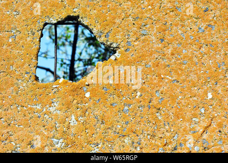 Yellow rough concrele wall with hole, blue sky and branches silhouette inside, surface close up detail, grunge horizontal shabby background Stock Photo