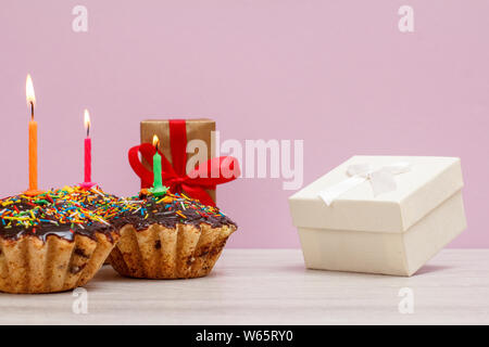 Gift boxes and three tasty birthday muffins with chocolate glaze and caramel, decorated with burning festive candles on the pink background. Happy bir Stock Photo