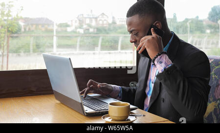 Afro american businessman working on laptop and talking mobile phone in cafe. He has a cup of coffee. Black man calling mobile phone and looking documents on computer. He wears shirt and suit jacket. Stock Photo