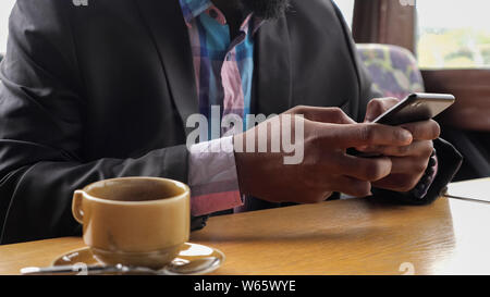 Afro american businessman is browsing smartphone sitting in cafe and drinking coffee, hands closeup. unrecognizable guy is flipping internet pages online in social media in mobile phone. Stock Photo