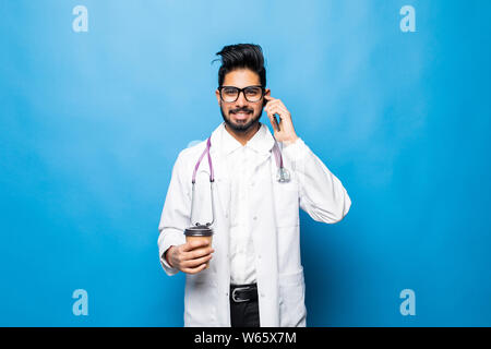Handsome indian doctor in white coat is using a smartphone, holding a cup of coffee and smiling while standing on blue background Stock Photo