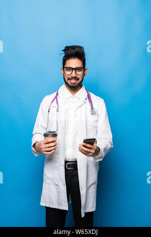 Handsome indian doctor in white coat is using a smartphone, holding a cup of coffee and smiling while standing on blue background Stock Photo