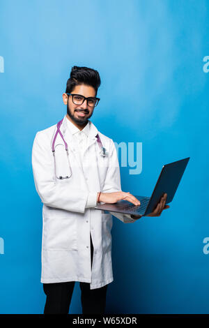 Indian doctor standing in the studio while working with laptop, isolated on blue background Stock Photo