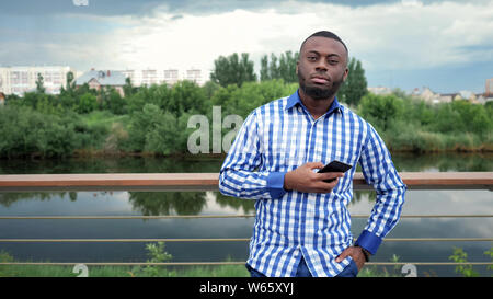 Young man browses phone looks at camera, copy space. Afro american guy smiles stands near fence on river waterfront in city park. He wears plaid shirt and jeans. Stock Photo