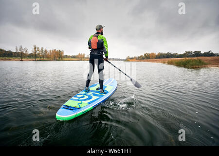 Athlete in wetsuit on paddleboard exploring the lake at cold weather against overcast sky Stock Photo