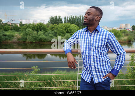 Afro american guy stands near fence on river waterfront in city park, copyspace. He wears plaid shirt and jeans. Stock Photo