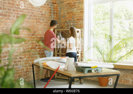 Young couple doing apartment repair together themselves. Married man and woman doing home makeover or renovation. Concept of relations, family, love. Put new furniture and books on the shelves. Stock Photo