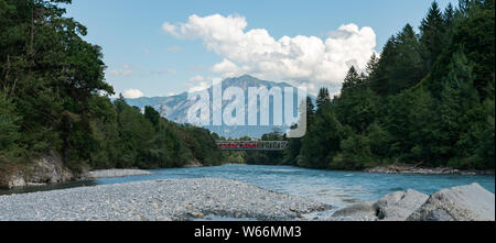 A red train crosses a bridge over the river Rhine in the Swiss Alps Stock Photo