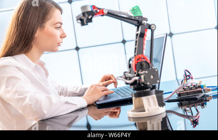 A young woman writes an algorithm for the robot arm. Science Research Laboratory for Robotic Arm Model. Computer Laboratory Stock Photo