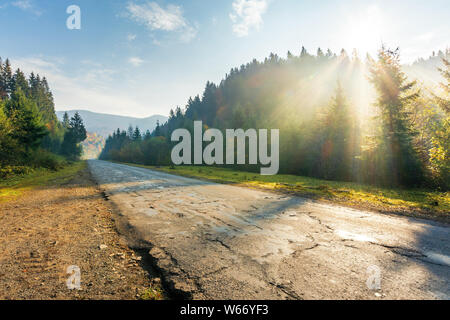 old road through forest in mountains at sunrise. beautiful transportation scenery in autumn. fluffy clouds on the azure sky. cracked asphalt and grave Stock Photo