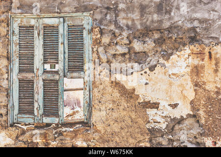 Old wooden window painted blue with broken shutters on the dirty cracked wall of a ruined house. Stock Photo