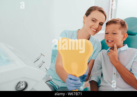 Little patient and his dentist smiling after successful dental treatment. They looking into mirror on perfect healthy teeth