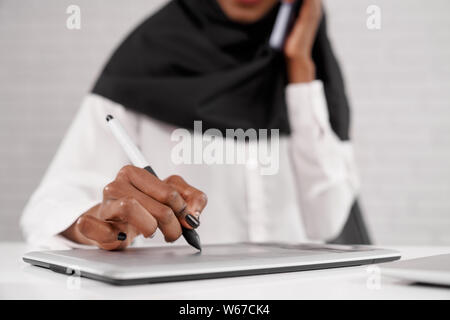 Selective focus of african woman keeping pen and writing on tablet while working. Young female sitting in office, talking by phone and communicating in company. Concept of technology and success. Stock Photo