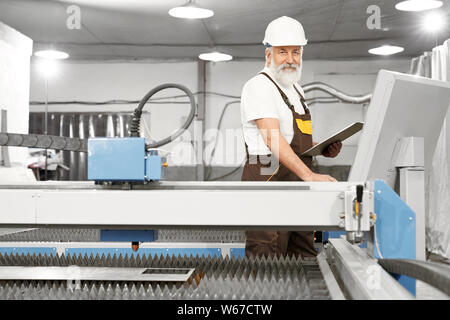Front view of smiling male engineer looking at camera and posing while working with computer on metal factory. Mechanic in uniform and helmet checking devices and writing data. Concept of industry. Stock Photo
