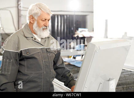 View from side of elder man wearing grey uniform working on factory. Bearded serious male worker looking at computer while using plasma cutter. Concept of engineering and manufacturing. Stock Photo