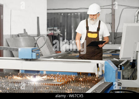 Professional engineer in uniform and helmet controlling process of plasma cutting on metal factory. Elder man keeping folded and checking devices and manufacturing. Concept of cnc and metalwork. Stock Photo