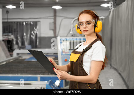 Side view of attractive female mechanic in uniform and protective headphones keeping folder and writing data on metal factory. Young woman looking at camera and posing. Concept of metalwork. Stock Photo