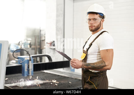 Side view of strong mechanic in protective glasses, helmet and uniform looking at camera and posing while working on metal factory. Male engineer standing near cnc and controlling plasma cutting. Stock Photo