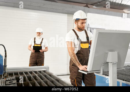 Two male engineers in helmets and uniforms in process of working on metal factory. Older man keeping folder and controlling cnc while mechanic using computer. Concept of machinery. Stock Photo