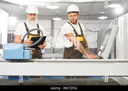 Front view of two skilled mechanics wearing uniform and helmets working together on metal factory. Experienced engineers standing near computer and cnc and controlling process of work. Stock Photo