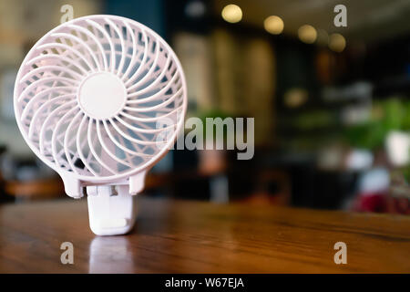 Close up of a portable battery operated fan, folded on a wooden table. The inner part is spinning with motion blur. There is a soft focus restaurant b Stock Photo