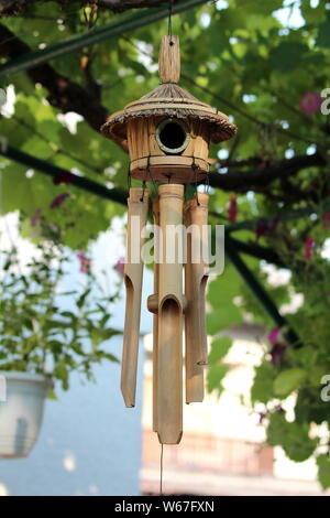 Garden decoration wooden wind chimes with small birdhouse on top surrounded with flowers and other plants in background on warm sunny spring day Stock Photo