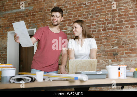Young couple doing apartment repair together themselves. Married man and woman doing home makeover or renovation. Concept of relations, family, love. Discuss the design of the wall using notebook. Stock Photo