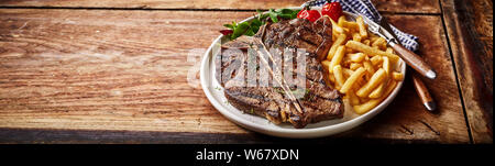 Panorama banner with barbecued T-bone steak and golden fried potato chips, French fries or pommes frites on an old rustic table with copy space Stock Photo