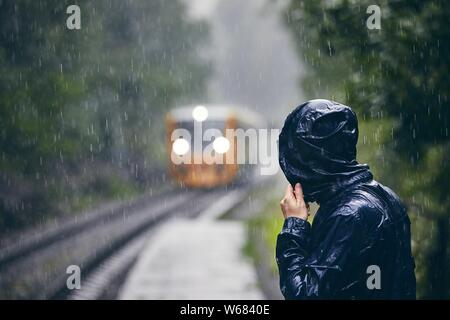 Man in drenched jacket standing on platform of railway station against train in heavy rain. Stock Photo