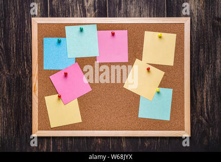 corkboard with pinned multicolored stickers on dark wooden background. Multicolor blank post it notes on a cork notice board. Stock Photo