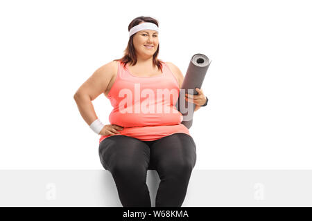 Plus size young female with an exercise mat sitting on a panel isolated on white background Stock Photo